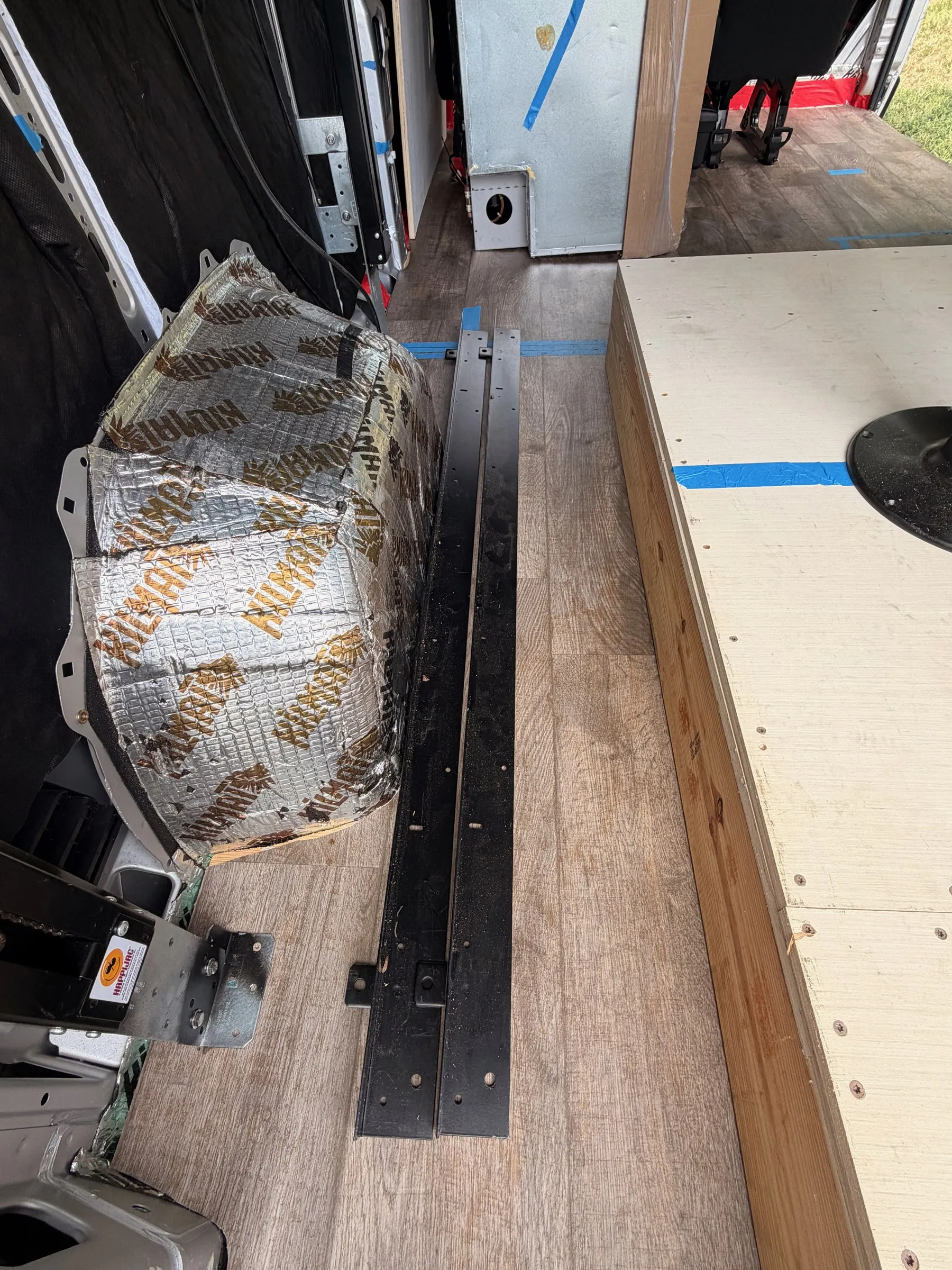 Wheel well wrapped in foil sound-deadening mat beside black mounting rails on the plank floor